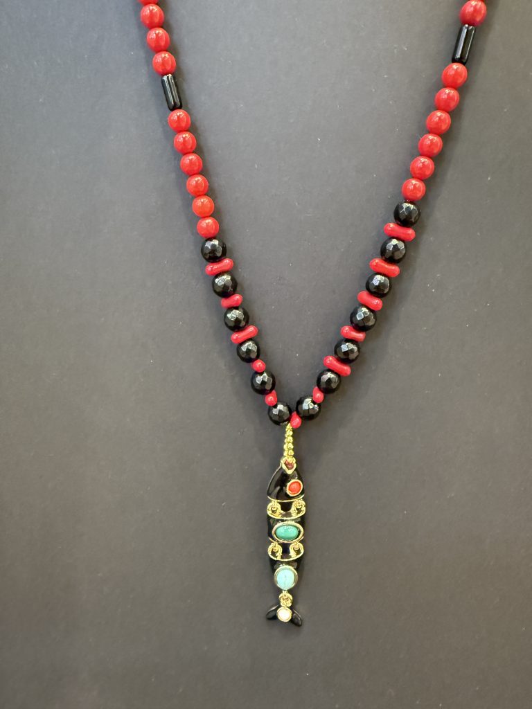 Μακρύ Vivid Tide Necklace (92εκ) από στρογγυλές κόκκινες χάντρες κοραλλιού και μαύρες χάντρες αχάτη, με χρυσά διαχωριστικά και ένα μεγάλο μεταλλικό στοιχείο σε σχήμα ψαριού διακοσμημένο με πολύχρωμες πέτρες.