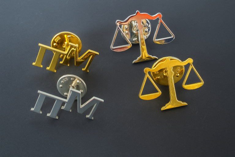Ποικιλία από custom pins (ασήμι 925) σε απλό φόντο, με δείγματα αρχικών γραμμάτων ('ΠΜ') και τη Ζυγαριά της Δικαιοσύνης. Αναδεικνύεται η δυνατότητα επιχρύσωσης και ο εξατομικευμένος σχεδιασμός.